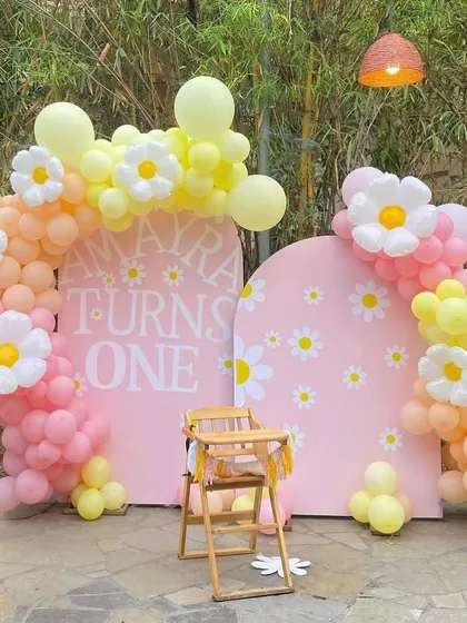 A sweet daisy-themed first birthday party with an outdoor feel. This decoration uses pink arched backdrops, pastel balloon garlands, and large daisy flower foil balloons, complemented by a wooden high chair for the baby.