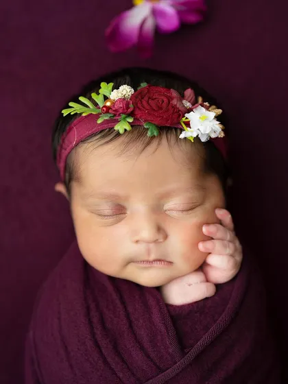 A close-up portrait focusing on the baby's serene expression and the beautiful floral headband that complements the deep plum wrap.