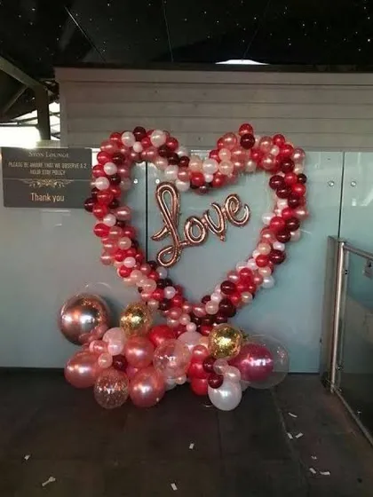 A heart-shaped balloon frame with a "Love" sign in the center, perfect for a Valentine's event entrance or photo spot. The base is decorated with metallic and clear balloons for extra sparkle.
