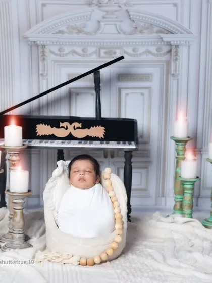A swaddled newborn rests in a basket in front of the piano. The warm glow from the candles creates a serene and classical atmosphere.