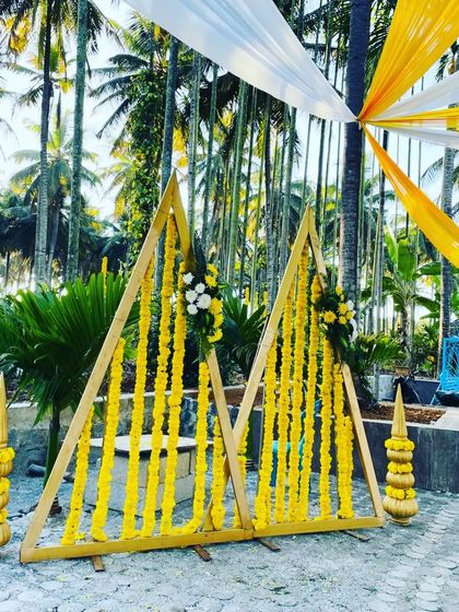 A wider shot of the triangular frames used for a haldi ceremony, showing the overhead canopy of yellow and white drapes. The setup includes traditional brass pots, blending modern and classic elements.