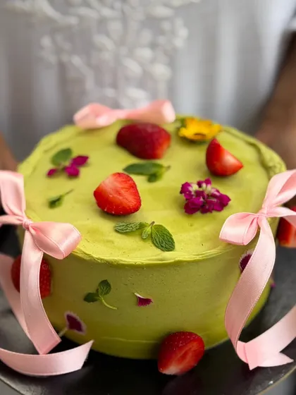 Holding up a finished custom Matcha Raspberry Cake, adorned with fresh strawberries, edible flowers, and delicate pink ribbons.