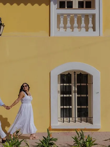 A candid walking shot against a vibrant yellow wall in Fontainhas, Goa. The bright colors and natural movement make this feel like a snapshot from a romantic holiday.