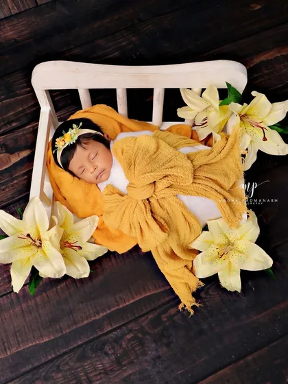 A baby wrapped in a yellow bow, resting on a white miniature bed and surrounded by beautiful yellow lilies.