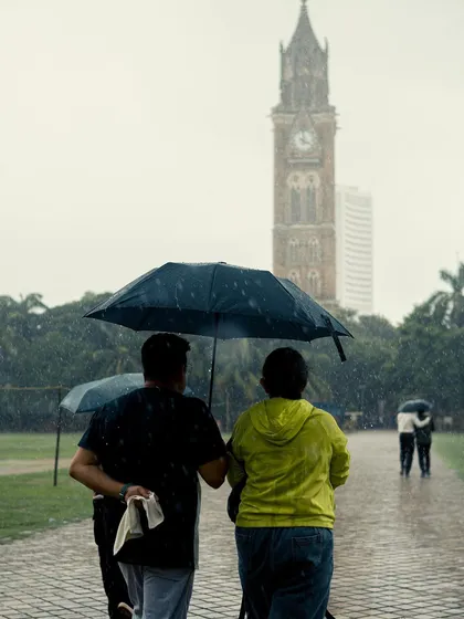 Two men walk together under an umbrella at Oval Maidan. It's a simple, candid shot of companionship, with the Rajabai Clock Tower providing a classic Mumbai backdrop.