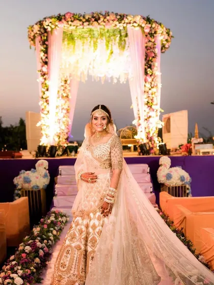 A stunning bride poses in front of her sunset wedding mandap. The decor features a mix of pink and white florals with cascading elements, creating a soft and romantic backdrop.