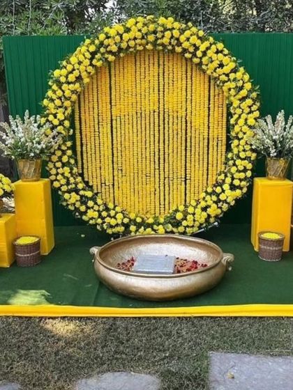 A classic Haldi backdrop for outdoor functions. This setup features a large circular frame filled with strings of yellow marigolds, a green panel background, and a traditional brass urli in the center for the ceremony.