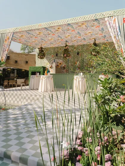 A wide view of a dreamy pastel garden party. The setup includes a custom bar with a floral canopy, a green and white checkered dance floor, and lush greenery, perfect for an elegant outdoor celebration.