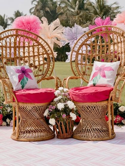 The bride's seating at a Goa Mehendi, featuring stylish peacock wicker chairs against a backdrop of giant, whimsical paper flowers. The vibrant pink cushions add a pop of color that ties the whole look together.