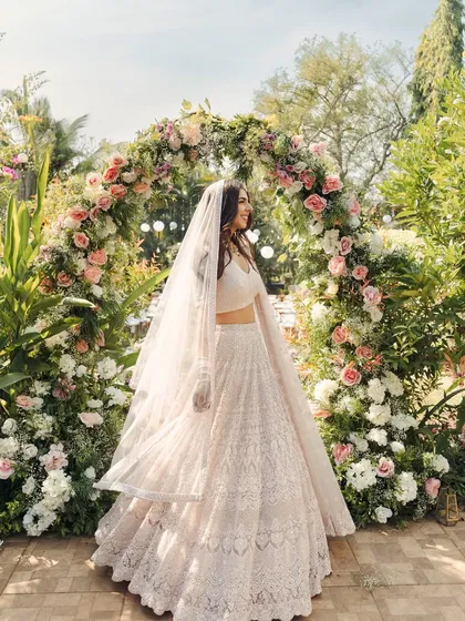 The bride, radiant in her all-white lehenga, stands before a magnificent floral arch. This portrait from her home garden wedding is full of light, joy, and natural beauty.