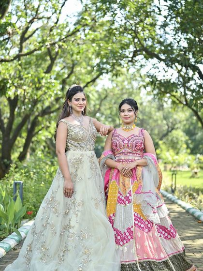 A lovely outdoor shot of two models. One in a pastel gown with a diamond necklace, the other in a colorful pink and yellow lehenga, perfect for a daytime event.
