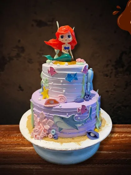 This two-tier mermaid cake features a beautiful purple and white design. It's decorated with edible shells, coral, and fish, with a lovely Ariel figure sitting on top.