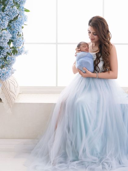 A mother in a stunning blue tulle gown holds her baby by a window, surrounded by blue flowers, creating a serene and color-coordinated portrait.