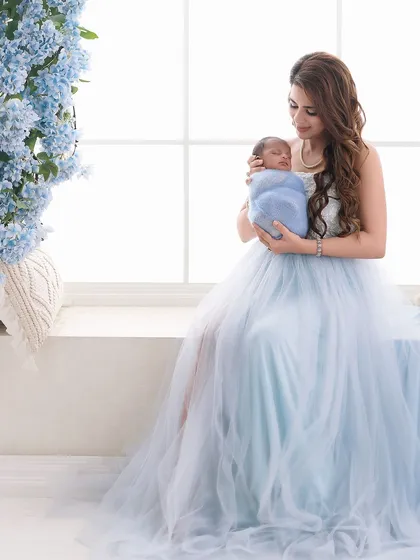 A full-length portrait of a mother in a flowing blue gown holding her swaddled newborn. The composition uses natural light and a hint of floral decor to create an elegant and dreamy image.