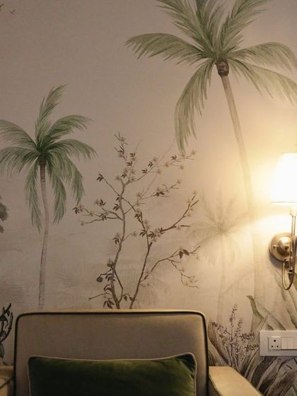 A closer view of the "whispering willow" wallpaper, where the warm glow of a wall lamp highlights the delicate details of the painted palm trees and foliage. This shows how lighting can interact with the art to create a specific mood.