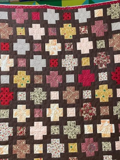A "plus" quilt with a warm, earthy color palette, set against a dark brown background.