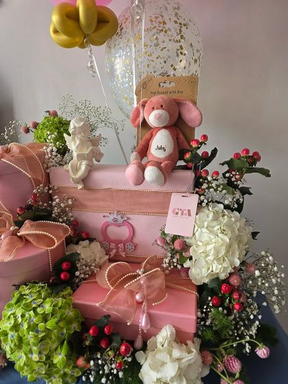 A beautiful baby girl hamper in shades of pink, featuring a plush bunny, angel figurines, and a lush arrangement of hydrangeas and berries.