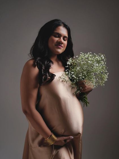 A soft and gentle portrait against a simple grey backdrop. The focus is on the mother's peaceful expression and the delicate baby's breath flowers.