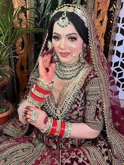 A beautiful portrait of a bride in a deep maroon velvet lehenga. The makeup features soft glittery eyes and a perfectly defined lip, complementing her traditional jewelry.