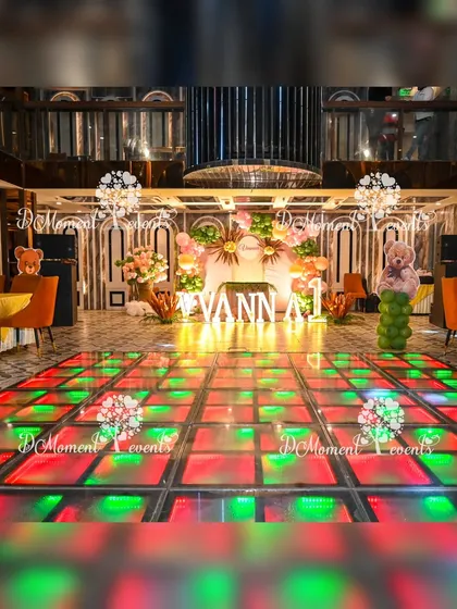 A wide shot of a teddy bear-themed party in a banquet hall with an LED dance floor. The stage is elegantly decorated with pastel balloons and boho elements.