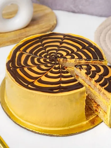 A full butterscotch cake with a chocolate web design, with one slice cut and ready to serve. This dynamic shot is perfect for showcasing whole cakes available for order.