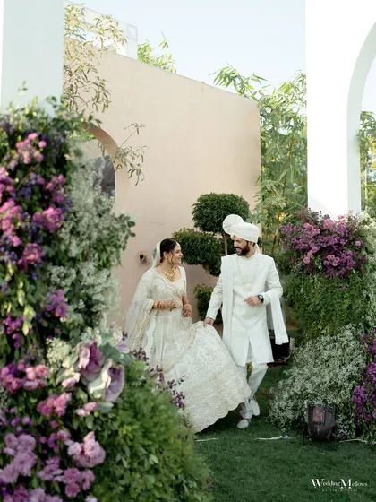 A candid shot of the couple walking through a garden, surrounded by beautiful floral arrangements. The bride's lehenga and the groom's sherwani are perfectly coordinated in elegant white.