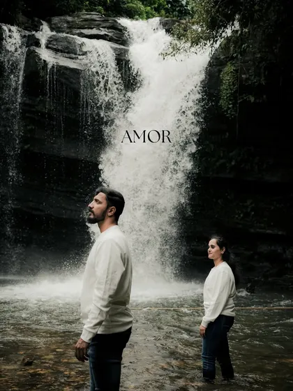An artistic and moody portrait of the couple standing before a powerful waterfall. The composition and the word "AMOR" create a strong, cinematic, and romantic statement.