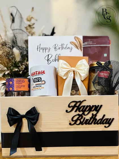 A birthday hamper for him, presented in a rustic wooden crate. It includes a "Vintage 1975" mug, gourmet coffee, and other treats, all styled with a masculine black and cream theme.