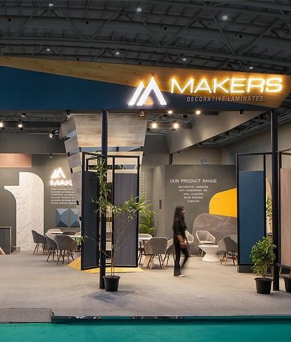 A full frontal view of the Makers Laminate exhibition stall. The open design allows clear sightlines into the booth, showcasing the central sculptural display and various meeting areas within.
