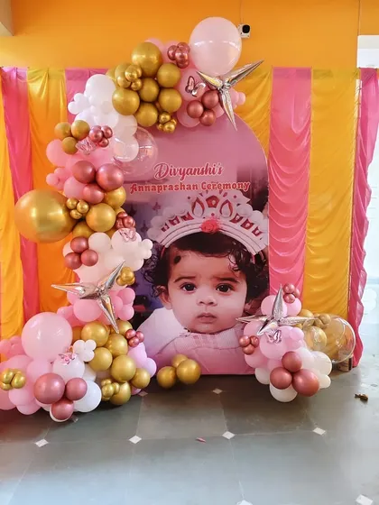 A personalized Annaprashan ceremony backdrop featuring a large photo of the baby. The arched backdrop is framed by an organic balloon garland in pink, rose gold, and white.