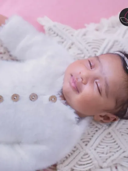A sleeping newborn with a gentle smile, dressed in a soft white outfit and adorned with a delicate floral headband.