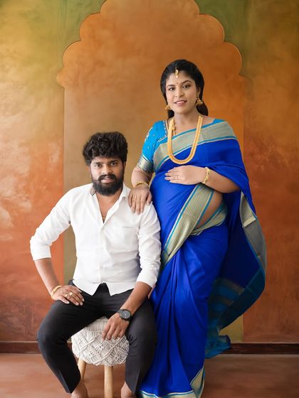 A strong and confident couple's portrait in a studio with a warm, textured backdrop. The mom-to-be glows in a vibrant blue saree.