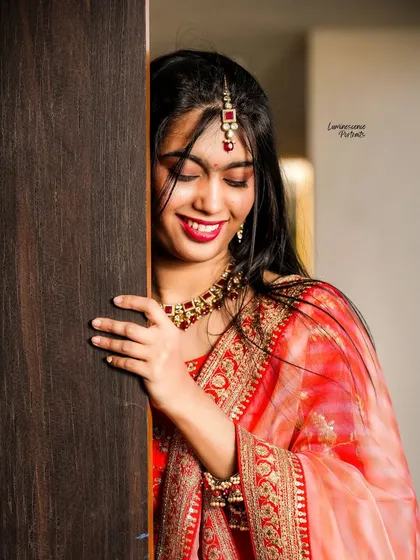 A shy, smiling bride peeks from behind a door, her red lehenga and jewelry glowing. A classic and sweet bridal portrait.
