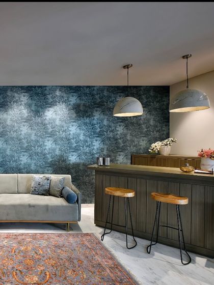 A cozy corner featuring a small bar setup. The deep teal wallpaper with a subtle pattern creates a moody backdrop, complemented by the warm wood of the bar and the soft grey of the sofa.