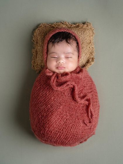 Simplicity is so beautiful. A close-up of a baby in a cozy, rust-colored wrap and bonnet, sleeping on a rustic burlap pillow. This highlights their perfect, tiny features.