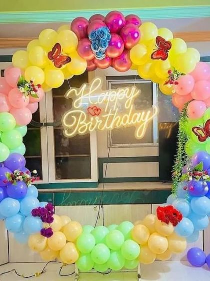 A bright and cheerful rainbow butterfly-themed birthday decoration. The circular balloon arch is adorned with small butterfly and flower details, all centered around a neon sign.