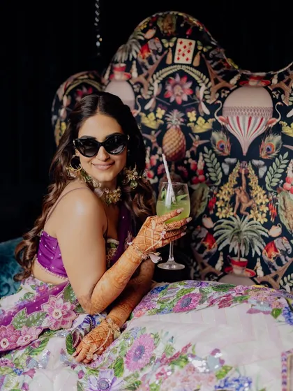 A modern bride at her Mehendi, looking cool and chic with sunglasses and a drink in hand. Her floral lehenga and the quirky printed chair create a stylish, relaxed vibe.