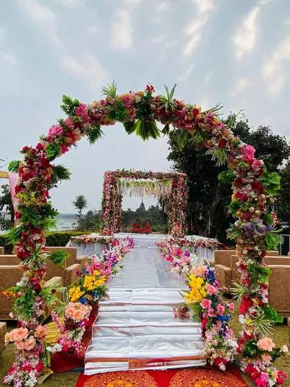 A breathtaking outdoor mandap with a full floral archway, set against a beautiful riverside view. The aisle is lined with colorful flowers.