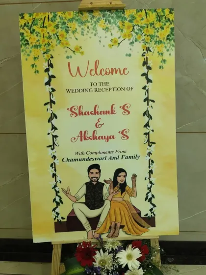 A personalized welcome sign for a wedding reception. The sign features a cute caricature of the couple and is decorated with a fresh flower arrangement at the base.