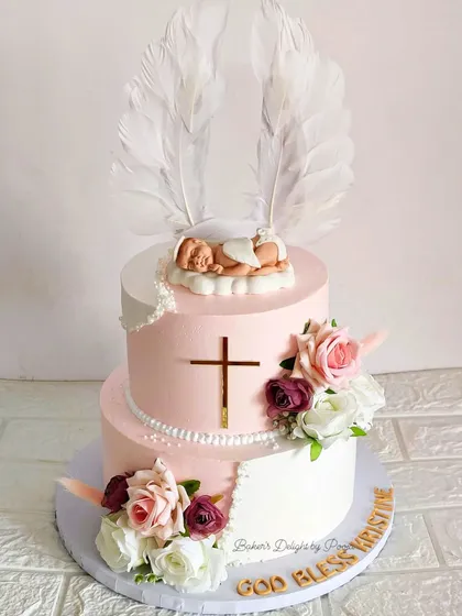 A serene and beautiful two-tier cake for a baby's baptism. The design features a soft pink and white palette, a sleeping baby angel topper with feather wings, fresh flowers, and a golden cross.