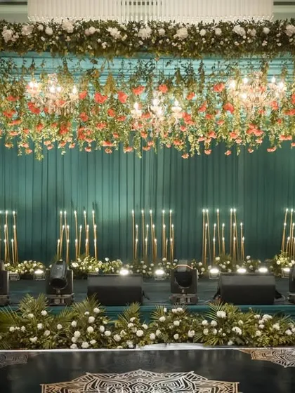 A luxurious stage design featuring a dramatic ceiling of hanging florals and chandeliers over a teal-paneled backdrop. The warm glow of candle-like floor lamps adds a layer of sophistication, ideal for a formal corporate dinner or awards ceremony.