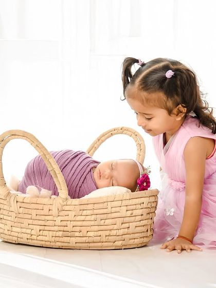 A curious older sister peeks into the basket at her sleeping newborn sibling. This candid moment, captured against a clean white background, is full of innocence and wonder.
