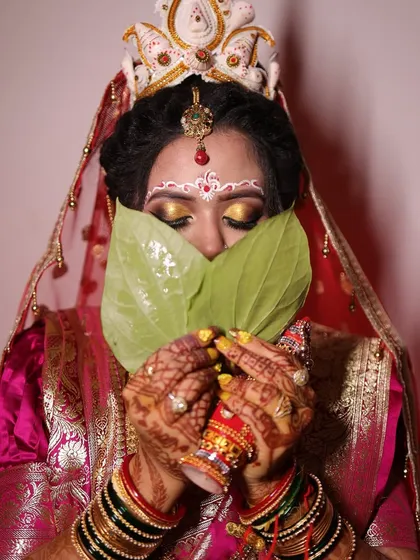 The iconic beauty of a Bengali bride, captured in the traditional 'Shubho Drishti' moment. The intricate white and red chandan designs on the forehead are a hallmark of this look, paired with golden eyeshadow and a classic red saree.