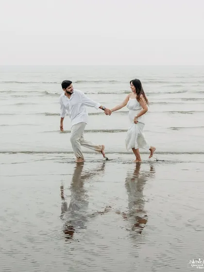 A classic romantic shot of a couple walking hand-in-hand in the shallow waters of a Mumbai beach. The reflection in the wet sand adds a beautiful, symmetrical element to the composition.
