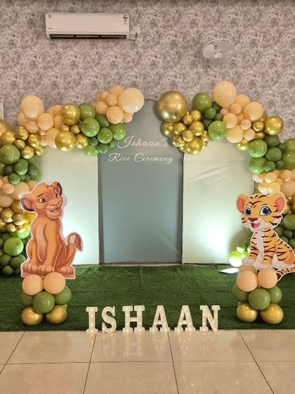 A sweet and simple jungle-themed rice ceremony decoration. The setup uses a green and gold balloon arch and cutouts of Simba and a tiger cub on an artificial grass base.
