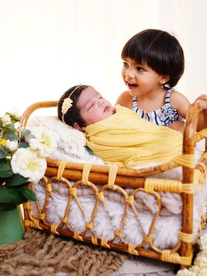 A beautiful moment between sisters, as the older sister looks lovingly at her newborn sibling sleeping in a small bed prop.