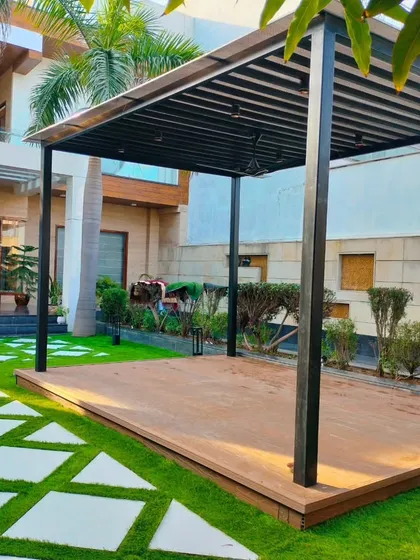 A front yard landscape design in Dwarka, New Delhi, featuring a modern metal pergola on a raised deck. The design uses geometric stone pathways set in lush artificial turf, demonstrating how we transform ground-level spaces in addition to rooftops.