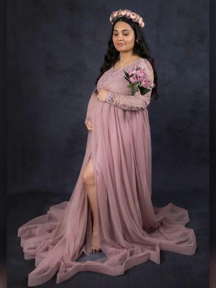 Fine art portraits can also be soft and romantic. This mother-to-be wears a dusty pink lace gown with a long train, complemented by a floral crown and a small bouquet for a touch of gentle elegance against a dark backdrop.