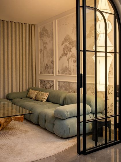 An intimate oasis of calm. This cozy nook features a plush, curved sofa set against a wall with nature-inspired art panels. The arched glass door adds a classic touch, separating the space while maintaining a visual connection.