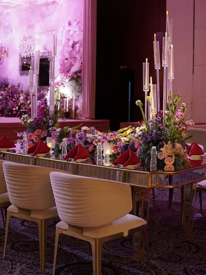 A luxurious dining table with a mirrored surface, set with a lush floral runner and tall candelabras, all under a ceiling of hanging crystal installations.